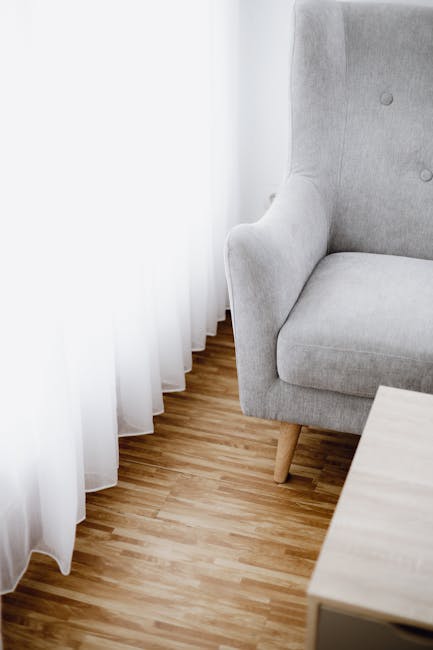 A close-up view of a corner in a residential living room, featuring a light grey upholstered armchair with wooden legs positioned near floor-to-ceiling white curtains. The curtains are made of lightwe