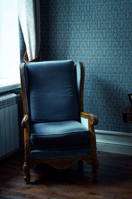A close-up view of a corner in a residential living room, featuring a light grey upholstered armchair with wooden legs positioned near floor-to-ceiling white curtains. The curtains are made of lightwe