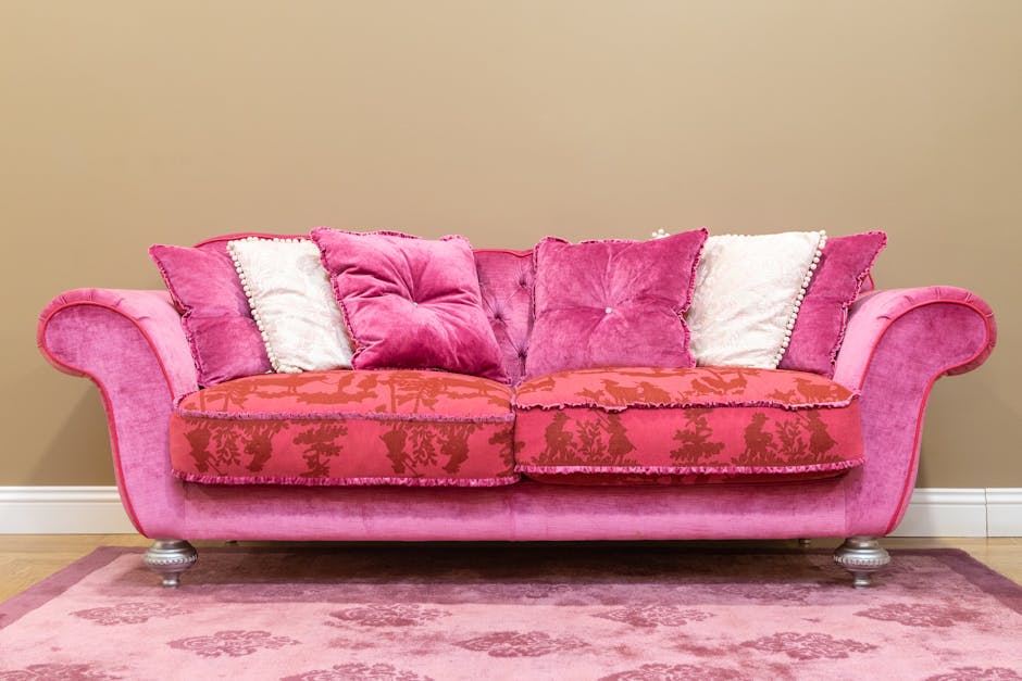 A close-up view of a light grey upholstered armchair with wooden legs, situated in a well-lit room with white curtains hanging over a window and a wooden floor. The armchair appears clean and free of 